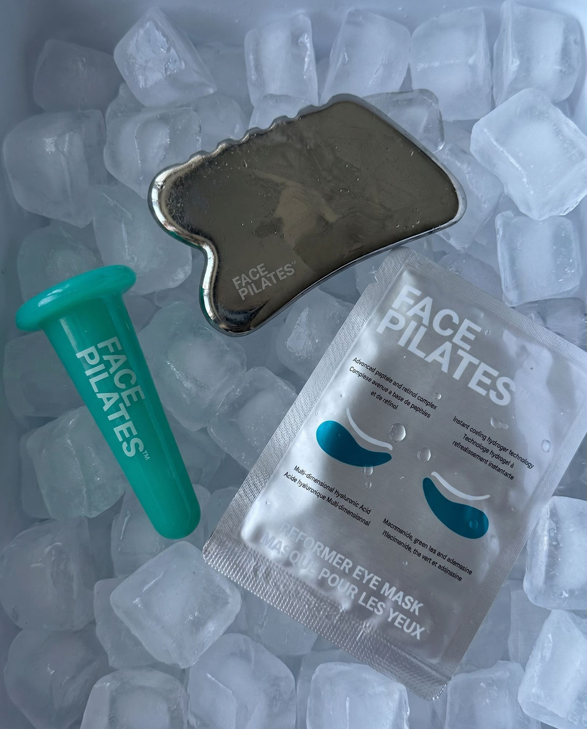 Face Pilates skincare set arranged on ice cubes, including a metallic forehead/eye massager tool, a teal applicator wand, and a packet of water-based eye mask patches.