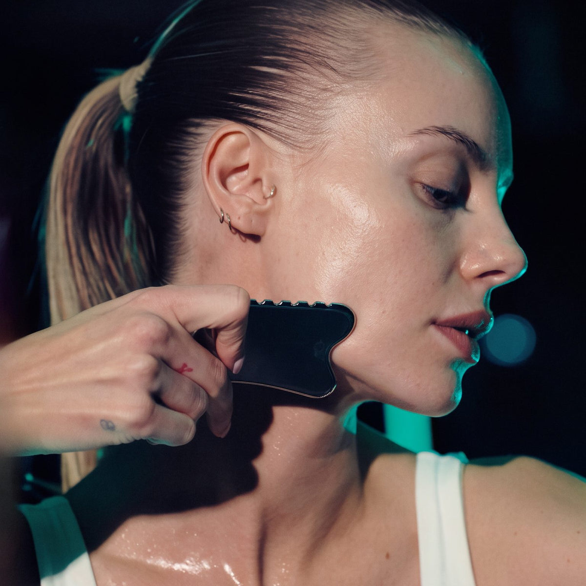 A woman in a white tank top has her hair pulled back and is shown from the side while another person uses a black facial scraping tool on her cheek and jawline. The scene is lit with dramatic teal and dark lighting against a dark background.