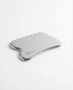 A polished stainless steel gua sha massage tool with a curved, scalloped edge and the 'FACE PLATES' logo printed on the side, shown on a clean white background.